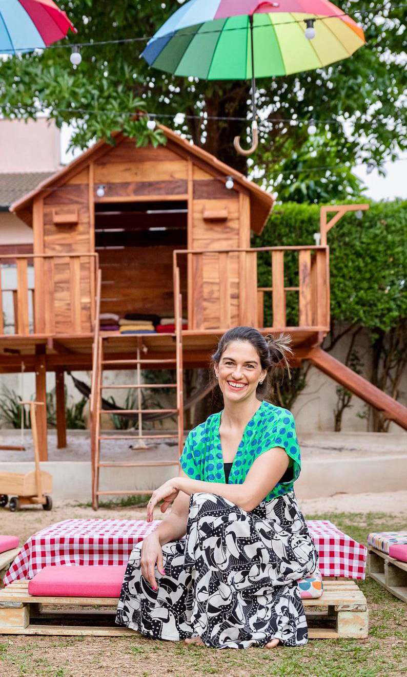 Carol Braga sentada no quintal, ao lado de uma casa de madeira